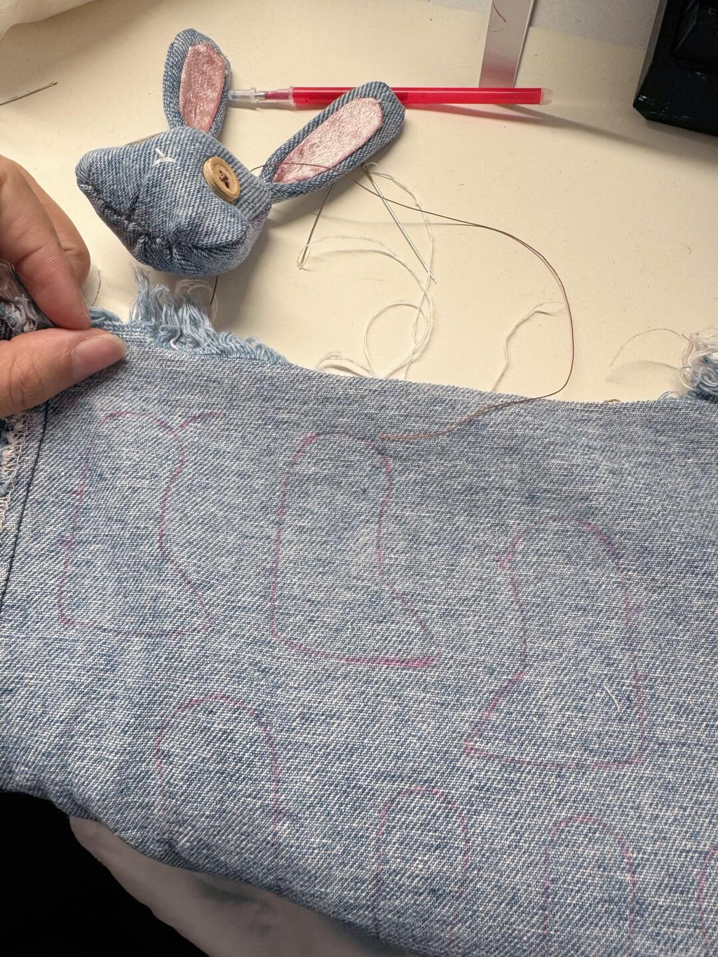 A close-up of a person holding a piece of light blue denim fabric with pink stitched bunny outlines, with a matching denim bunny head and pink inner ears visible in the background on a light tabletop.