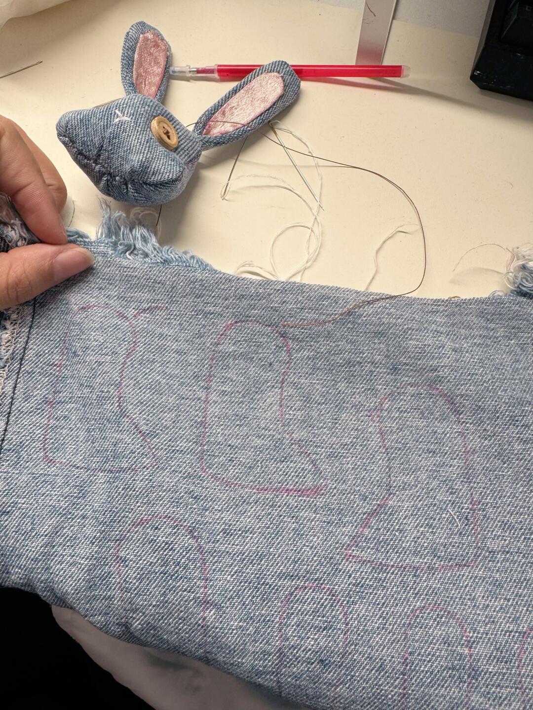 A close-up of a person holding a piece of light blue denim fabric with pink stitched bunny outlines, with a matching denim bunny head and pink inner ears visible in the background on a light tabletop.