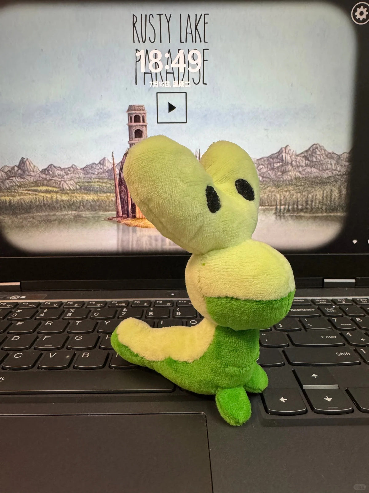 A green Applin and yellow plush toy resembling a cartoon creature sits on a black laptop keyboard, with the laptop screen showing a scenic landscape and the text 'RUSTY LAKE PARADISE' and a play button.