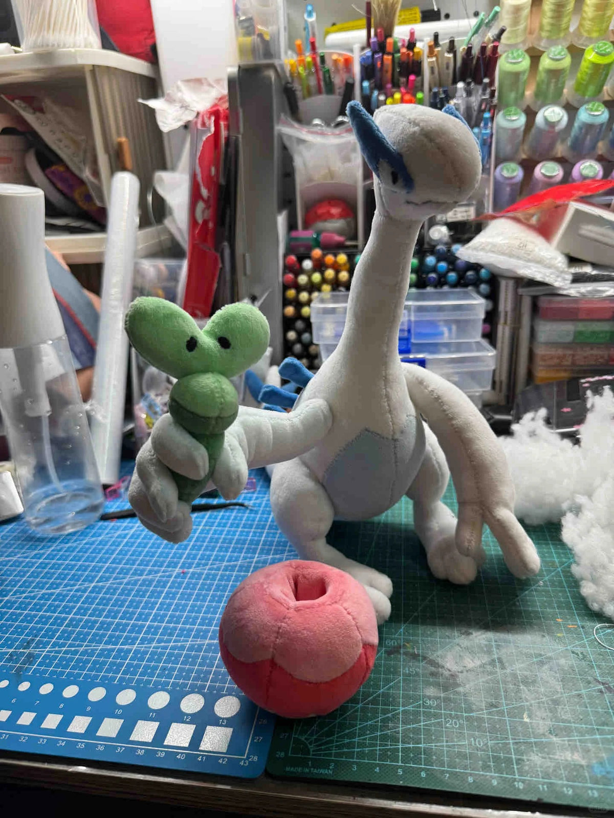 A handmade Applin  plush toy of a white bird-like creature with blue accents, holding a small green plush, with a pink and white plush ball in front, set on a blue cutting mat in a cluttered craft room.