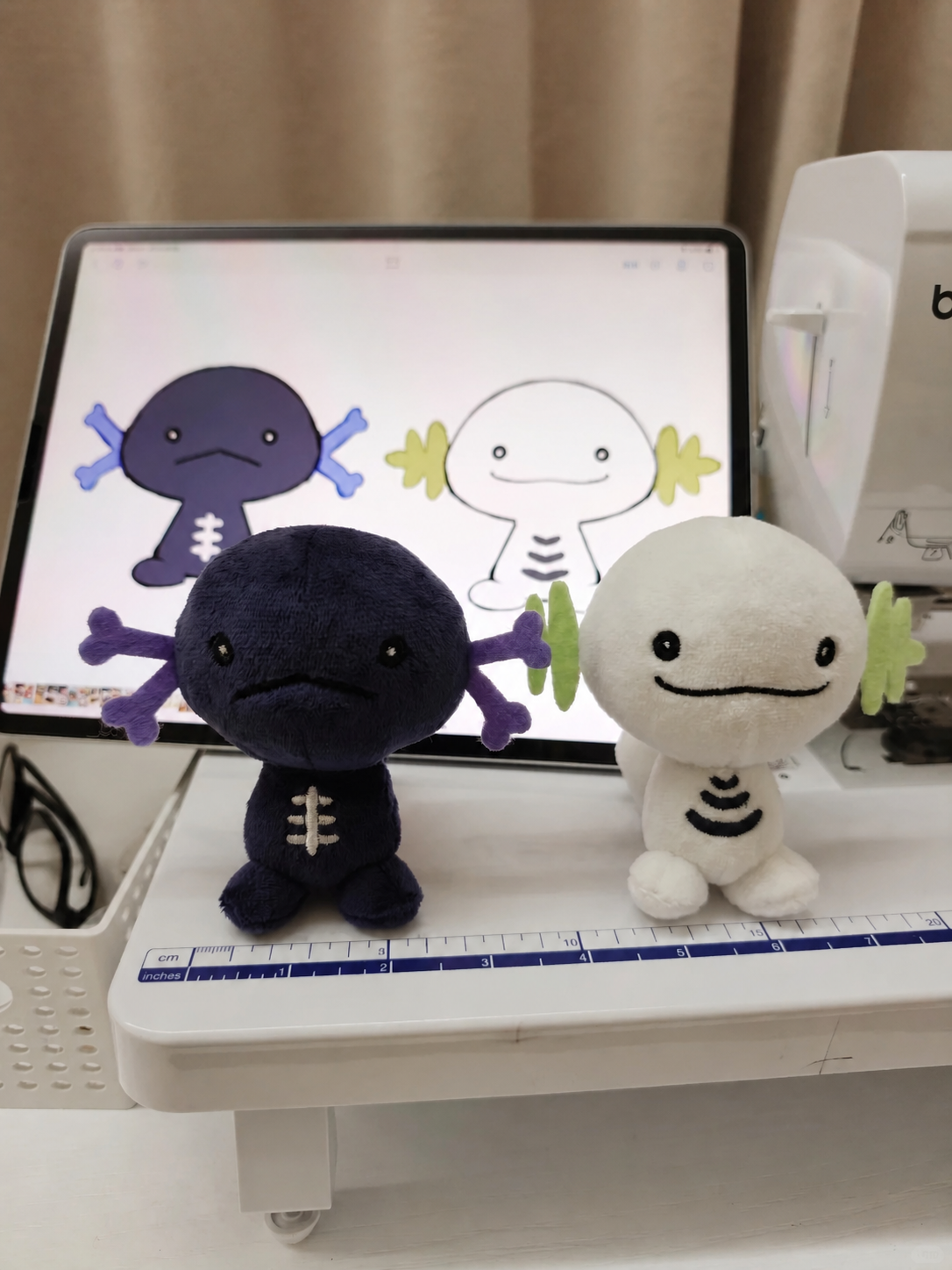 Two Wooper small plush axolotl toys displayed on a white table in front of a tablet showing digital designs of the same characters.