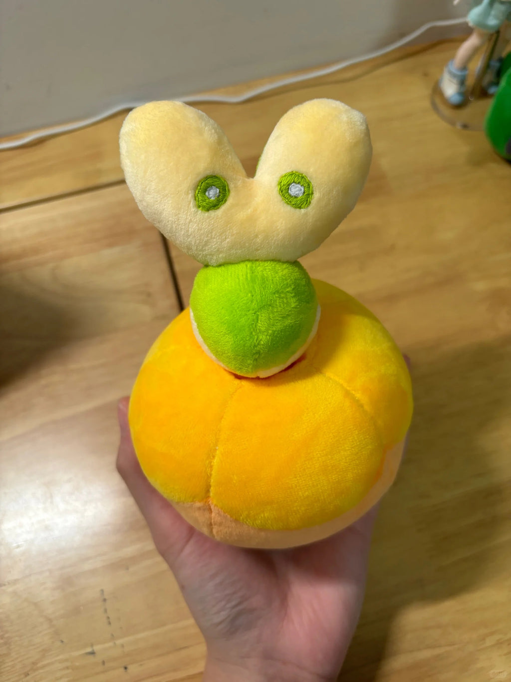 A small Applin plush toy held in a hand, featuring a round yellow-orange body, a green neck, and a pale yellow bunny head with green eyes.
