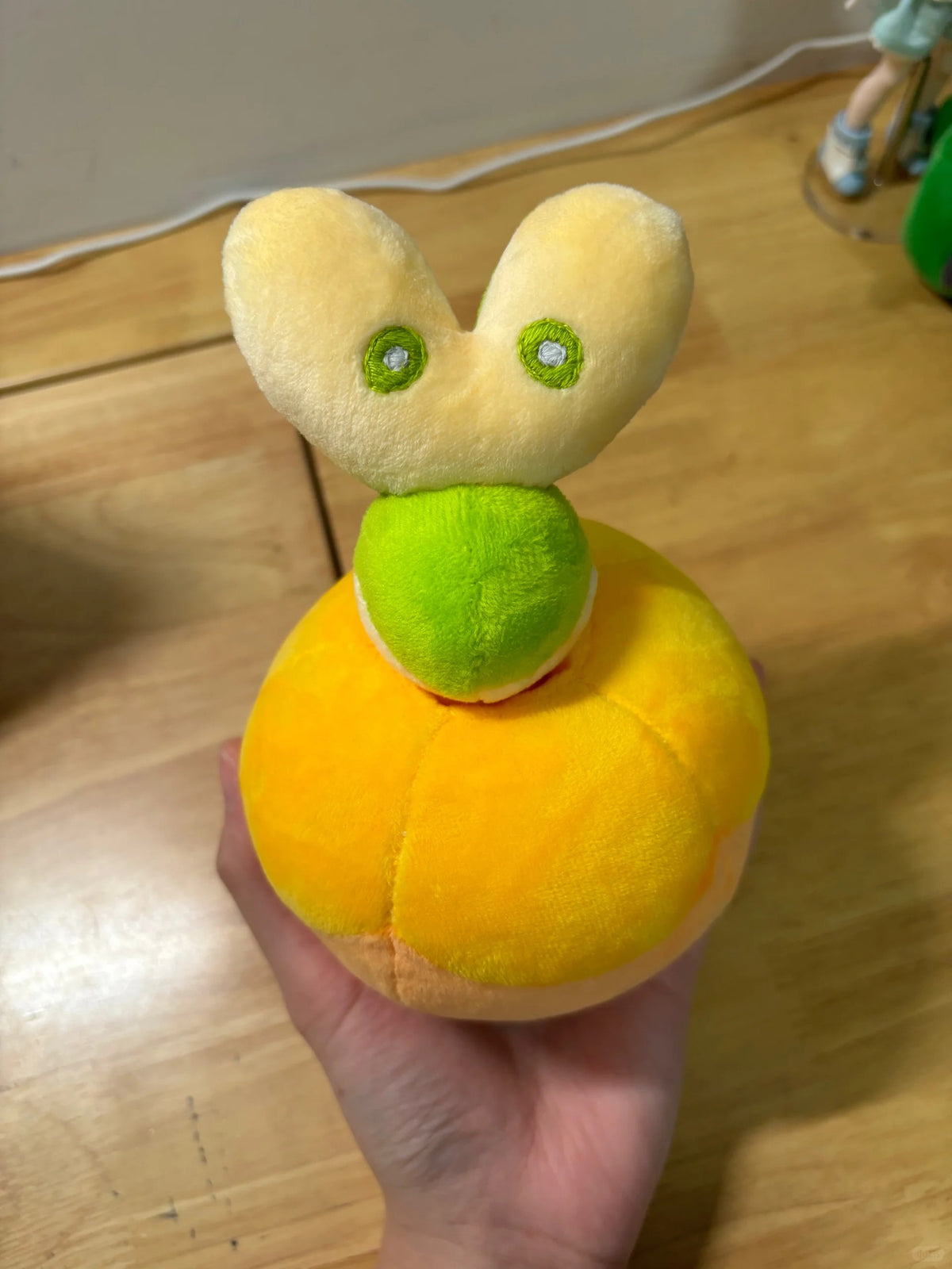 A small Applin plush toy held in a hand, featuring a round yellow-orange body, a green neck, and a pale yellow bunny head with green eyes.