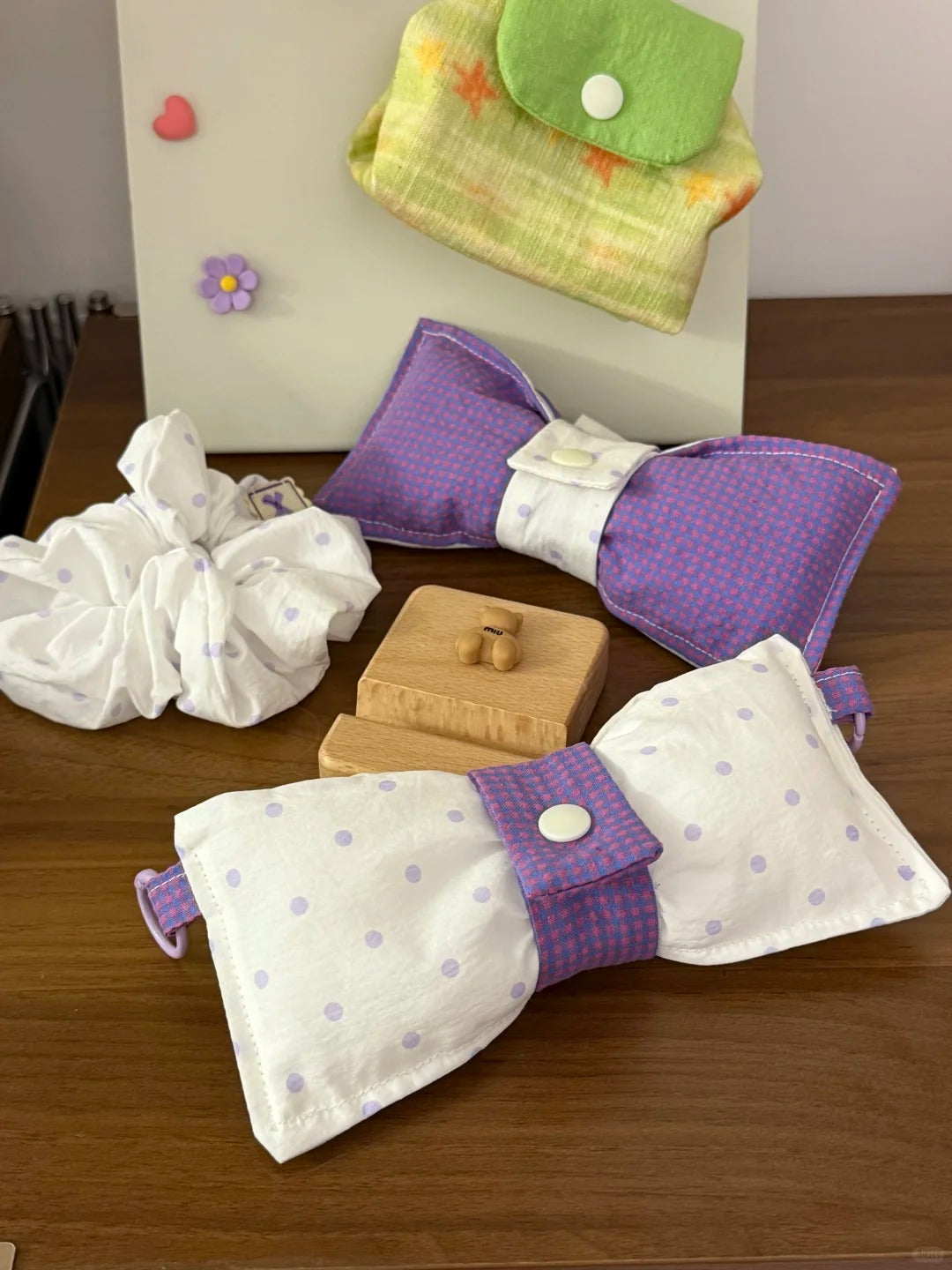 A set of handmade bow Glasses Pouches displayed on a wooden table, including two bow-shaped items in purple and white polka dot and solid purple fabrics, a white scrunchie, a small wooden block, and a green and yellow patterned pouch hanging on a white board in the background.