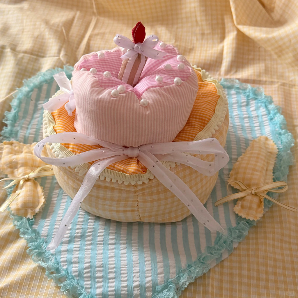 A handmade fabric baby keepsake gift set styled like a birthday cake, featuring a pink striped top, orange and cream gingham layers, a white ribbon bow, and a small red tassel candle, presented on a blue and yellow striped heart-shaped fabric mat.