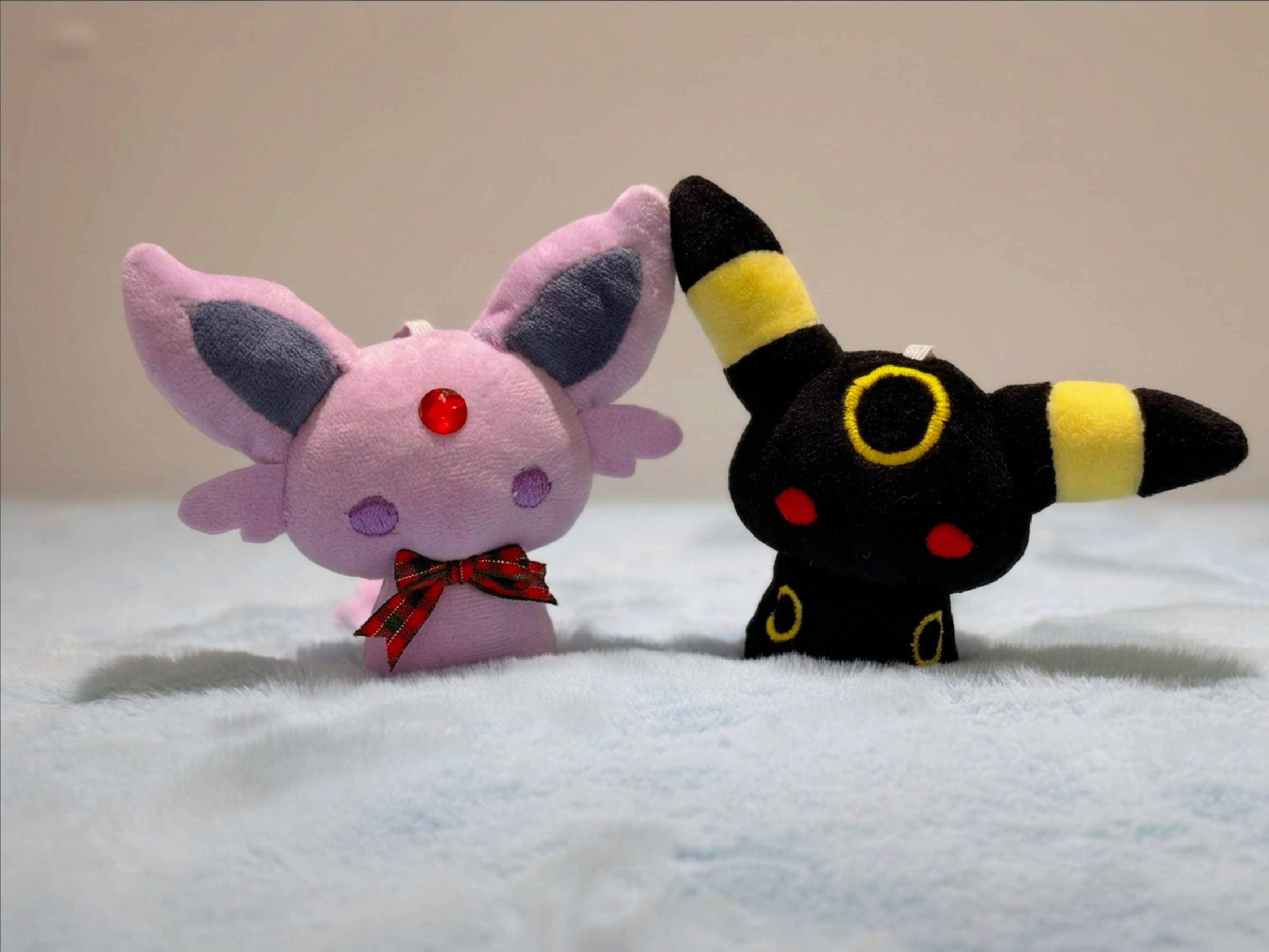 Two eevee evolutions plush toys, one pink with a bow tie and one black with yellow accents, on a light background.
