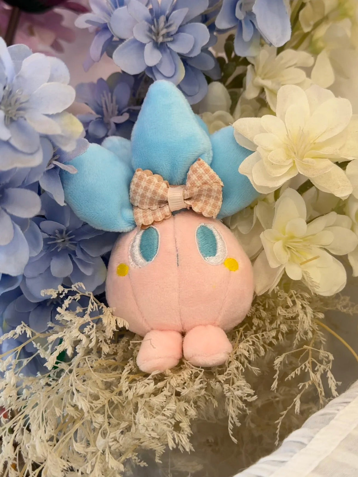 A cute pink oddish plush toy with blue wings and a checkered bow, nestled among blue and white artificial flowers.
