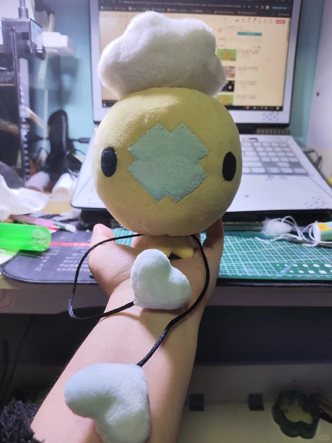 Yellow Drifloon plush colour variation held above a cutting mat and laptop