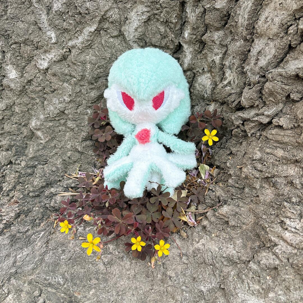 Handmade mint green Gardevoir plush doll displayed against tree bark and small yellow flowers