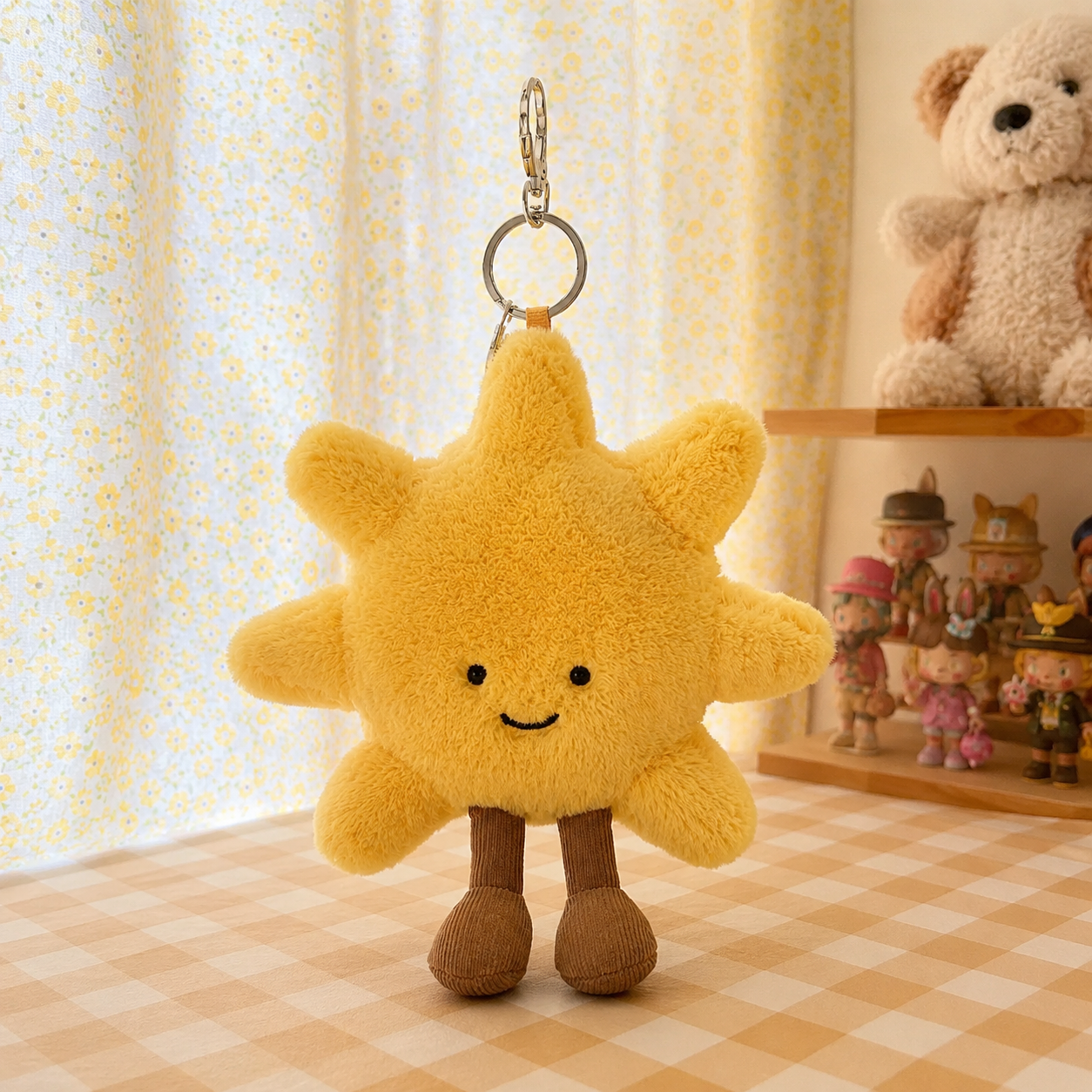 A yellow plush jellycat sun keychain with embroidered eyes and smile, standing on small brown legs in front of a yellow floral curtain, with a teddy bear and small figurines visible in the softly lit room background.