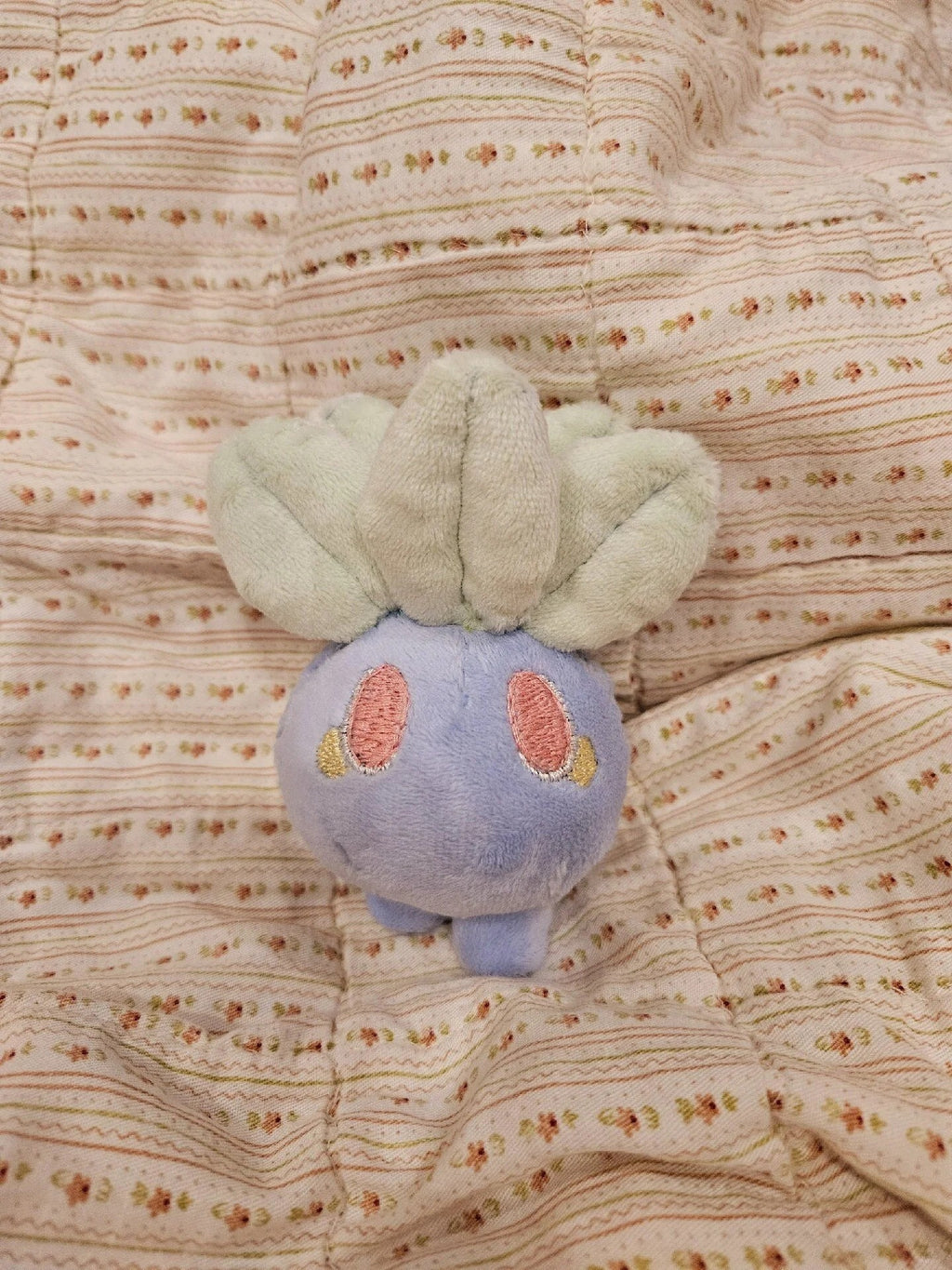 A small oddish plush toy resembling a cute blue creature with pale green leaf-like ears and pink cheeks, resting on a textured cream quilted blanket with fine floral stripes.