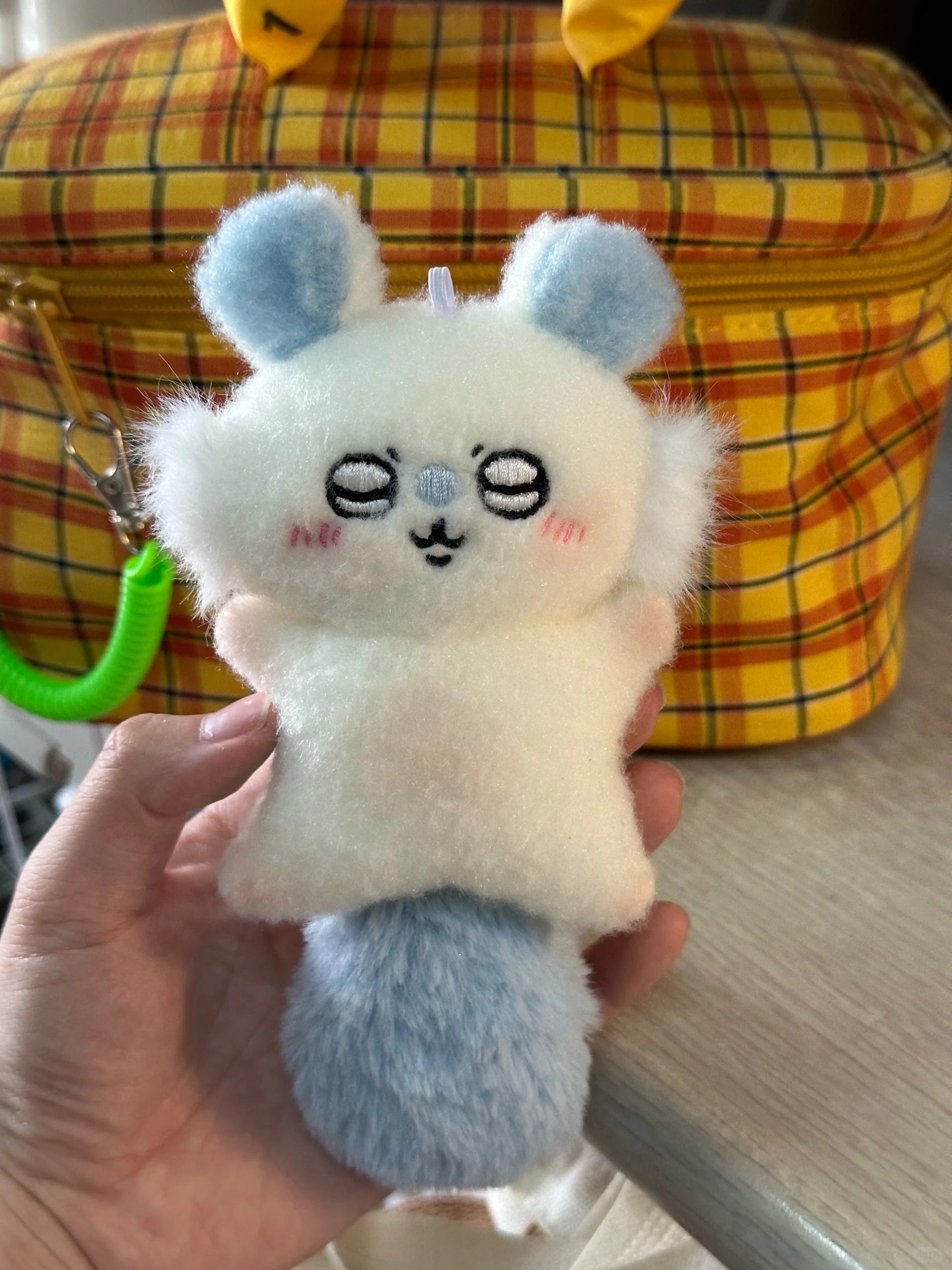 A hand holds a small momonga plush character keychain or bag charm in front of a yellow plaid bag. The plush is mostly white with light blue ears and tail, plus small pink cheeks and a simple embroidered face.