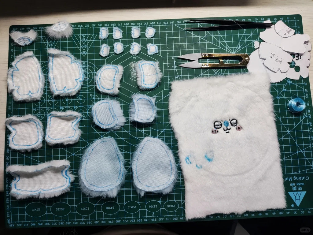 A set of handmade momonga plush toy or character body parts laid out on a green cutting mat, including white faux-fur pieces with blue stitched edges, a white furry panel with embroidered eyes and nose, small plastic cutout templates, tweezers, scissors, and a blue bead on a green grid cutting mat.