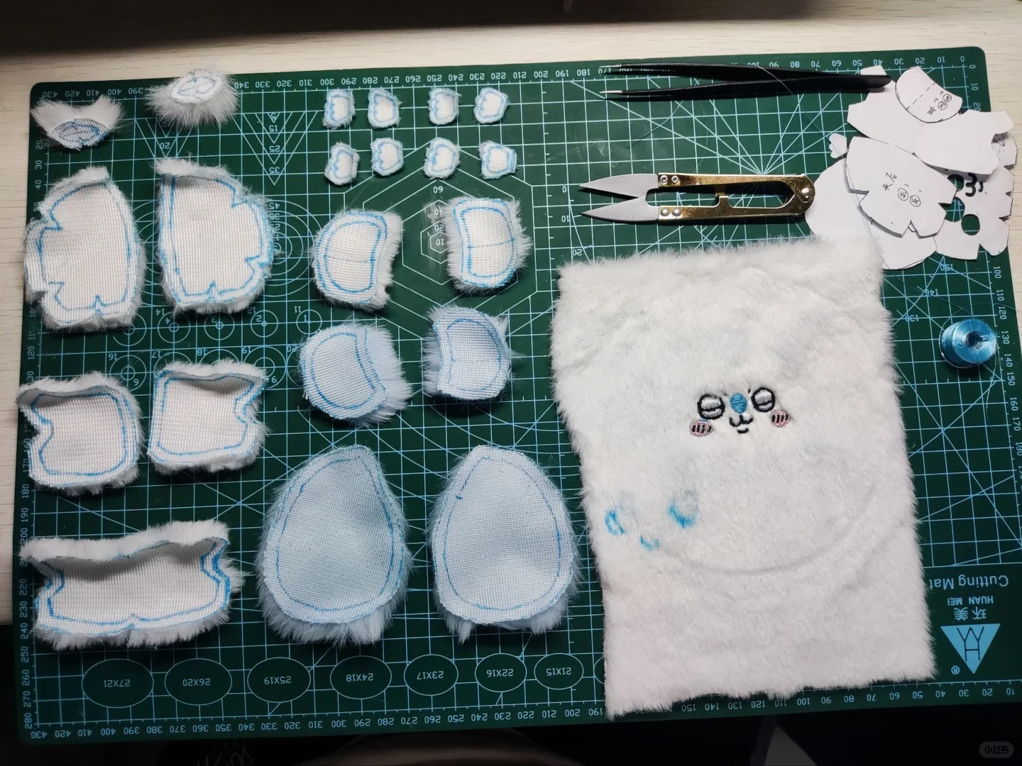A set of handmade momonga plush toy or character body parts laid out on a green cutting mat, including white faux-fur pieces with blue stitched edges, a white furry panel with embroidered eyes and nose, small plastic cutout templates, tweezers, scissors, and a blue bead on a green grid cutting mat.