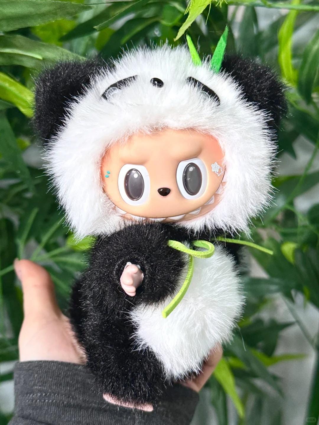 Plush toy resembling a panda with a labubu face held against a green leafy background