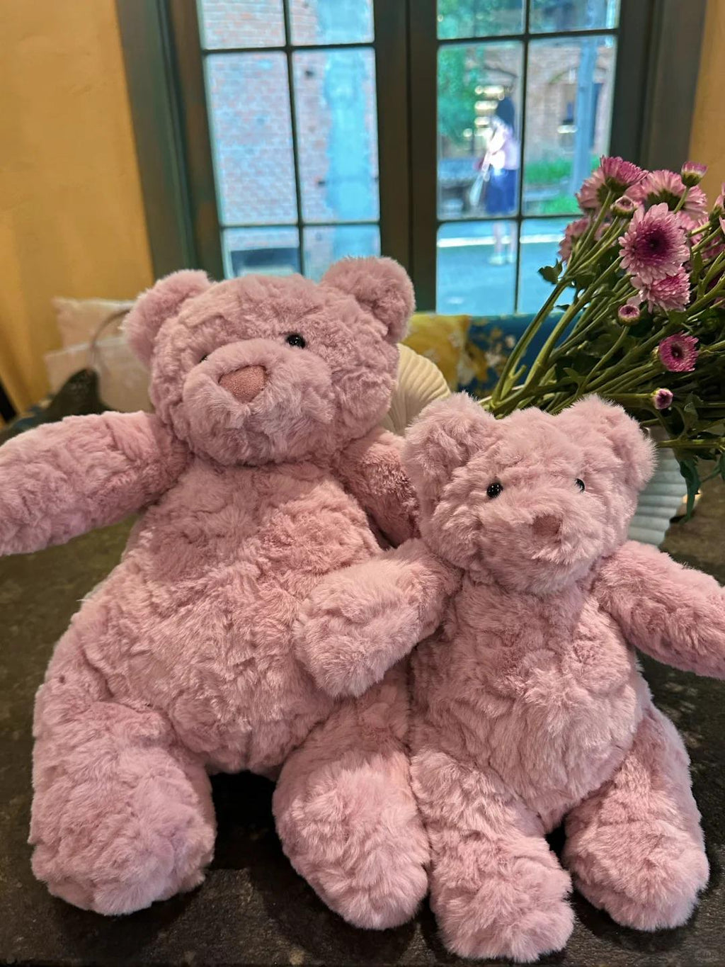 Two pink teddy bears sitting on a surface with flowers in the background.