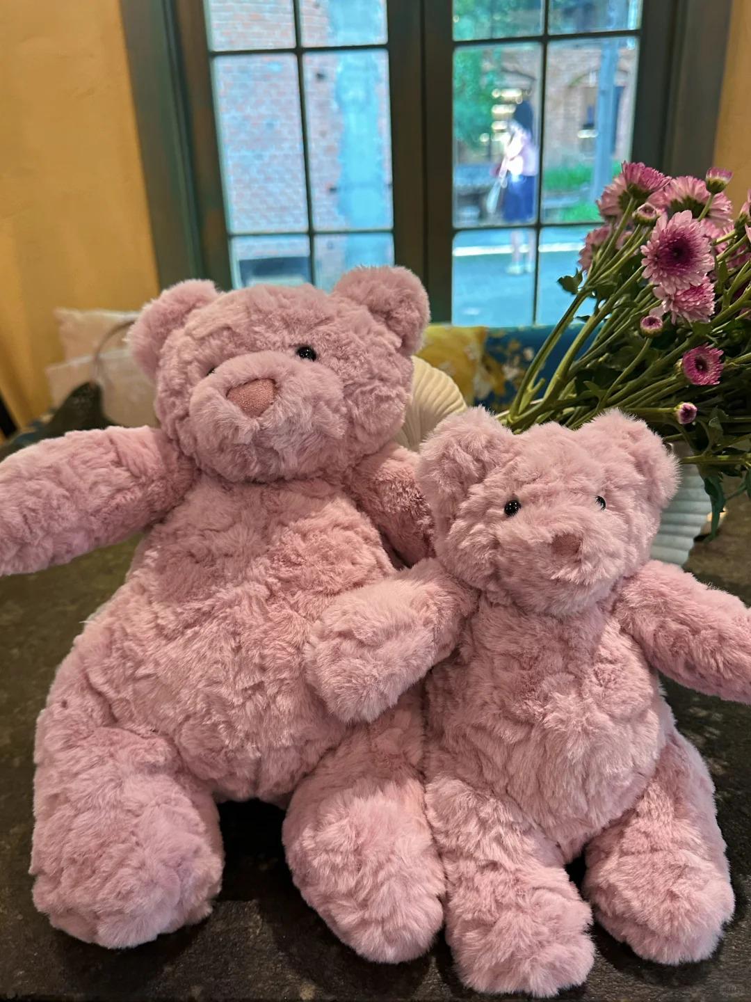Two pink teddy bears sitting on a surface with flowers in the background.