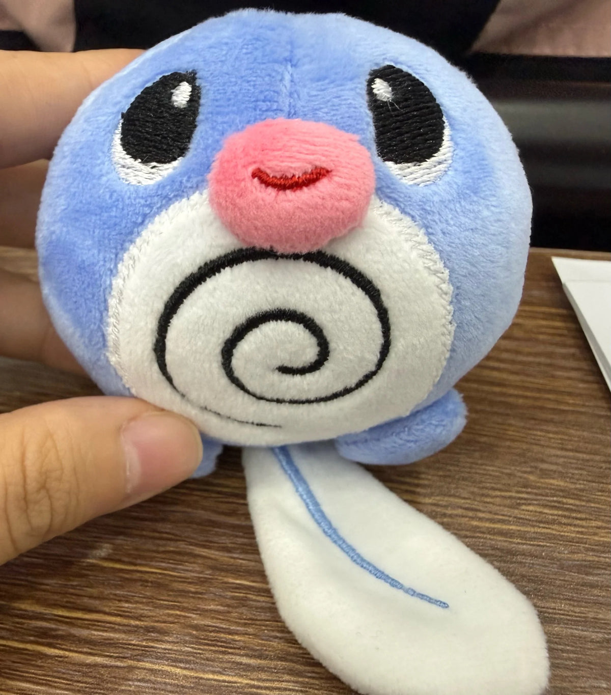 A small Poliwag plush toy resembling a blue and white cartoon creature with a pink nose, held in a person's hand over a wooden desk.
