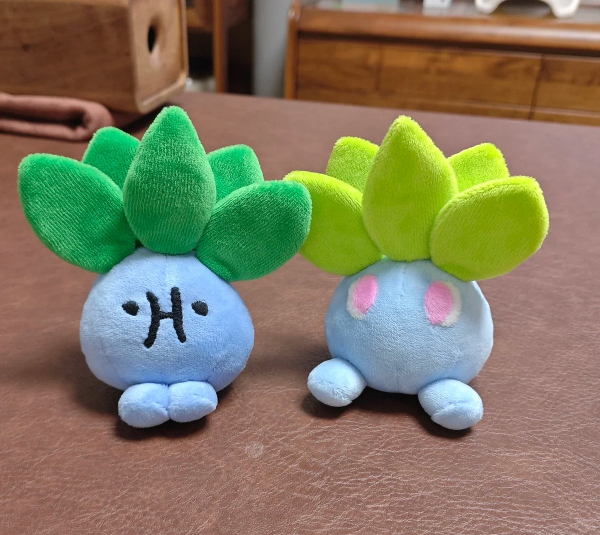 Two small oddish plush toy characters with light blue bodies and leaf-like tops, one in green and one in yellow-green, placed on a brown tabletop.