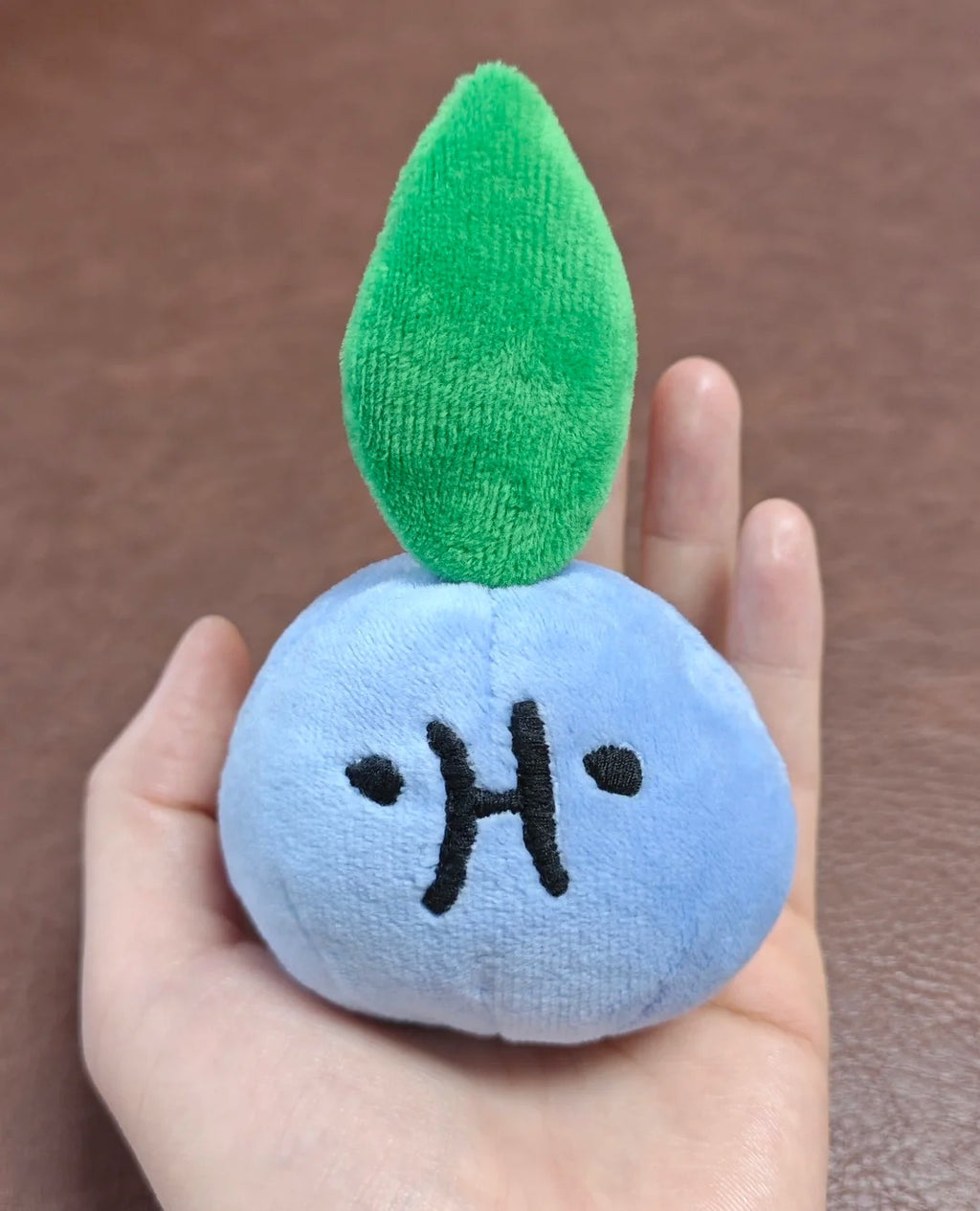 A small oddish plush toy shaped like a round light blue fruit or seed with a bright green leaf on top and a simple black embroidered face, being held in a person's hand against a brown textured background.