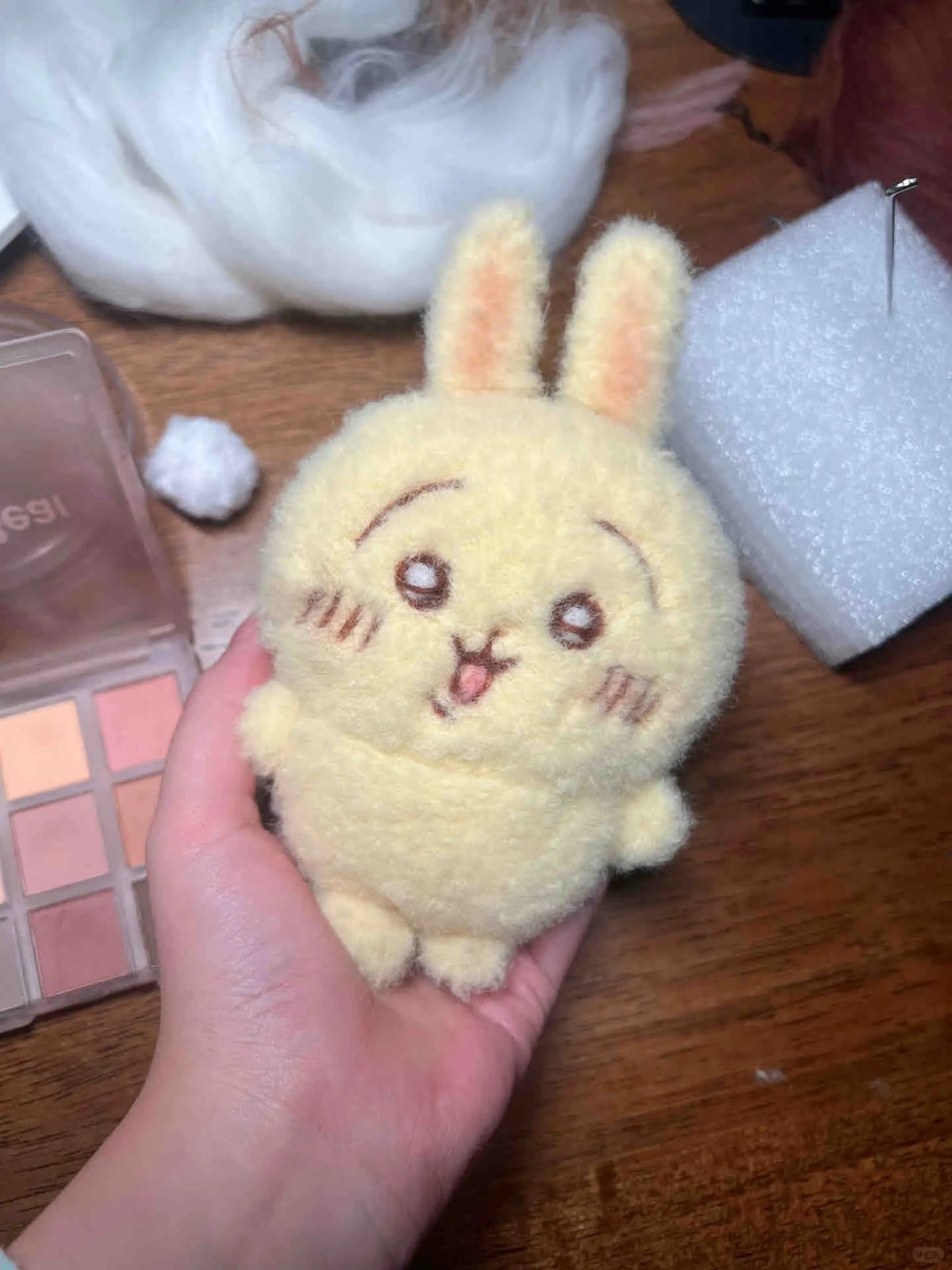 A hand holding a small fuzzy plush bunny character with long ears, embroidered facial features, and a pale yellow color, set against a wooden table with craft supplies.